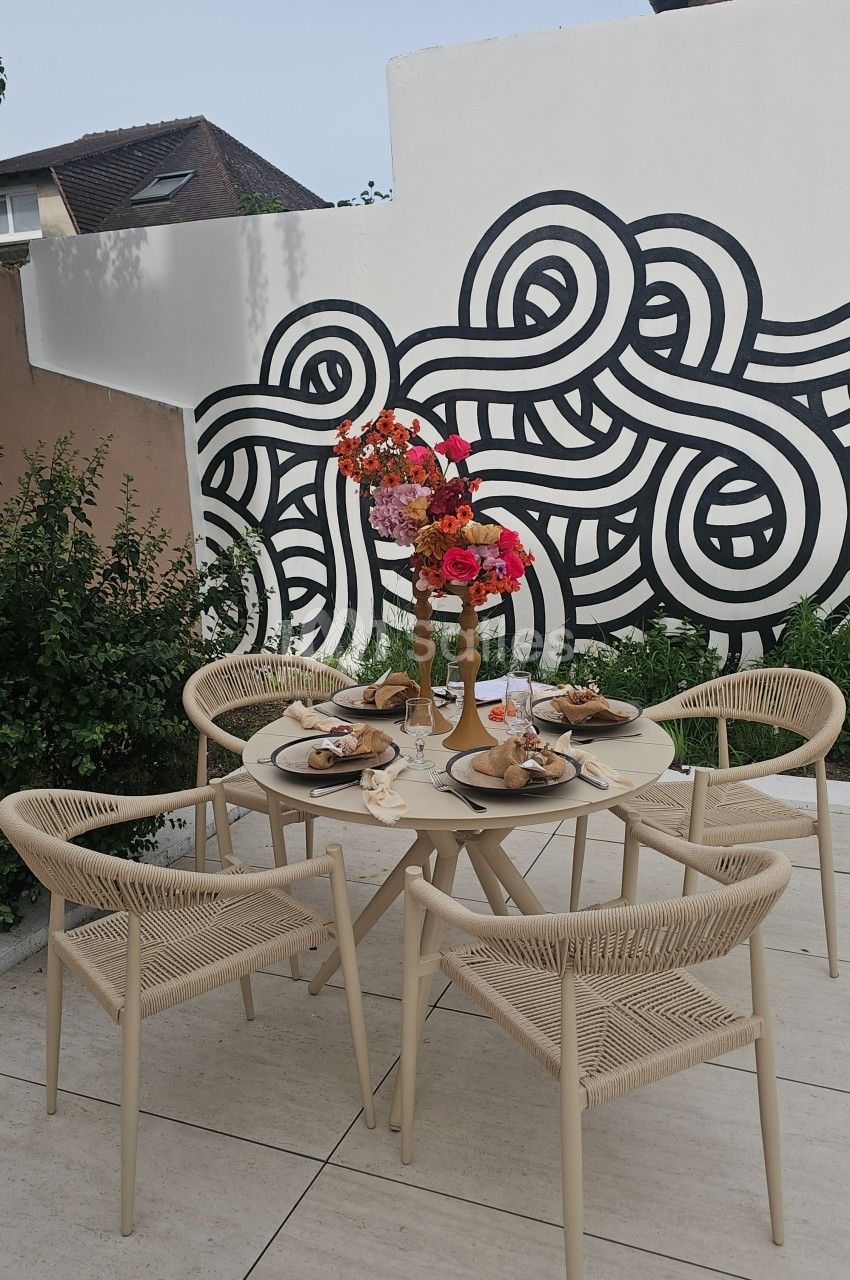 Table de jardin beige avec chaises assorties, décorée de fleurs colorées et dressée pour un repas, devant une fresque murale.