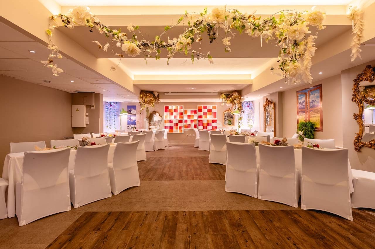 Salle de réception décorée avec des fleurs suspendues, des tables dressées et des chaises blanches alignées.