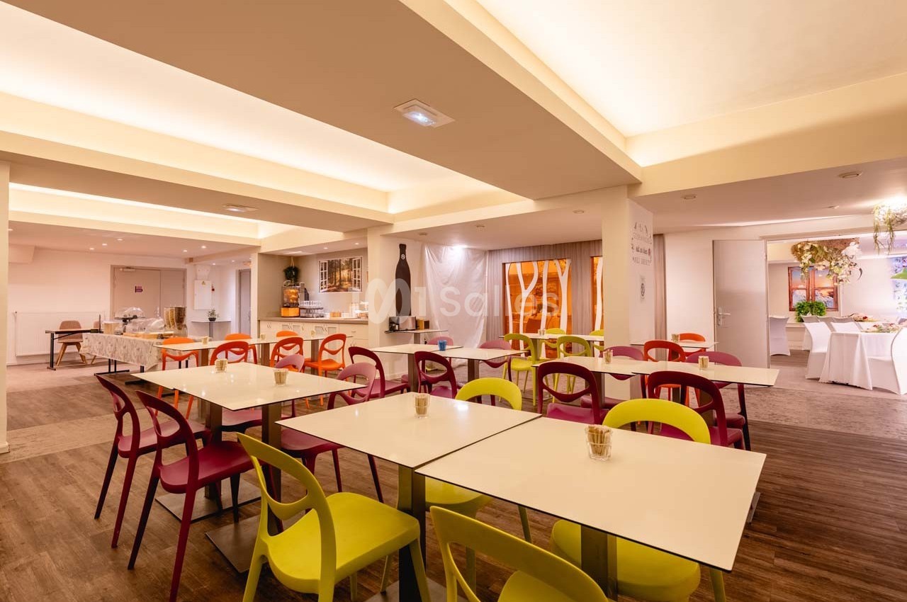 Salle lumineuse avec tables blanches, chaises colorées et buffet en arrière-plan, dans un espace moderne.