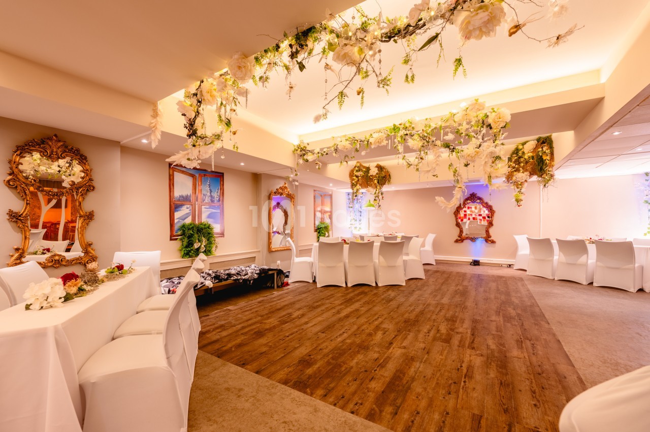 Salle décorée pour un événement, avec chaises blanches, tables dressées, guirlandes florales et miroirs dorés.