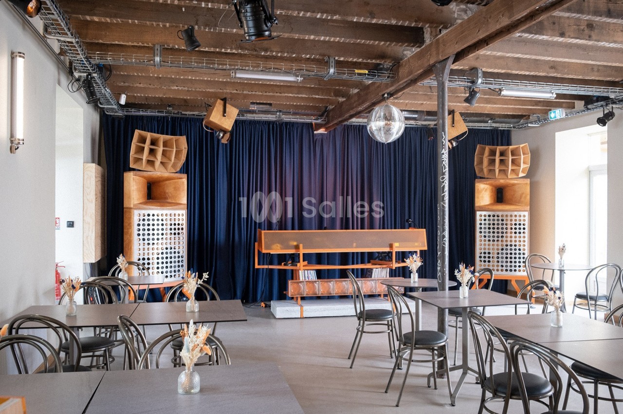Salle avec tables, chaises, scène équipée de matériel audio, rideaux bleus et plafond en poutres apparentes.