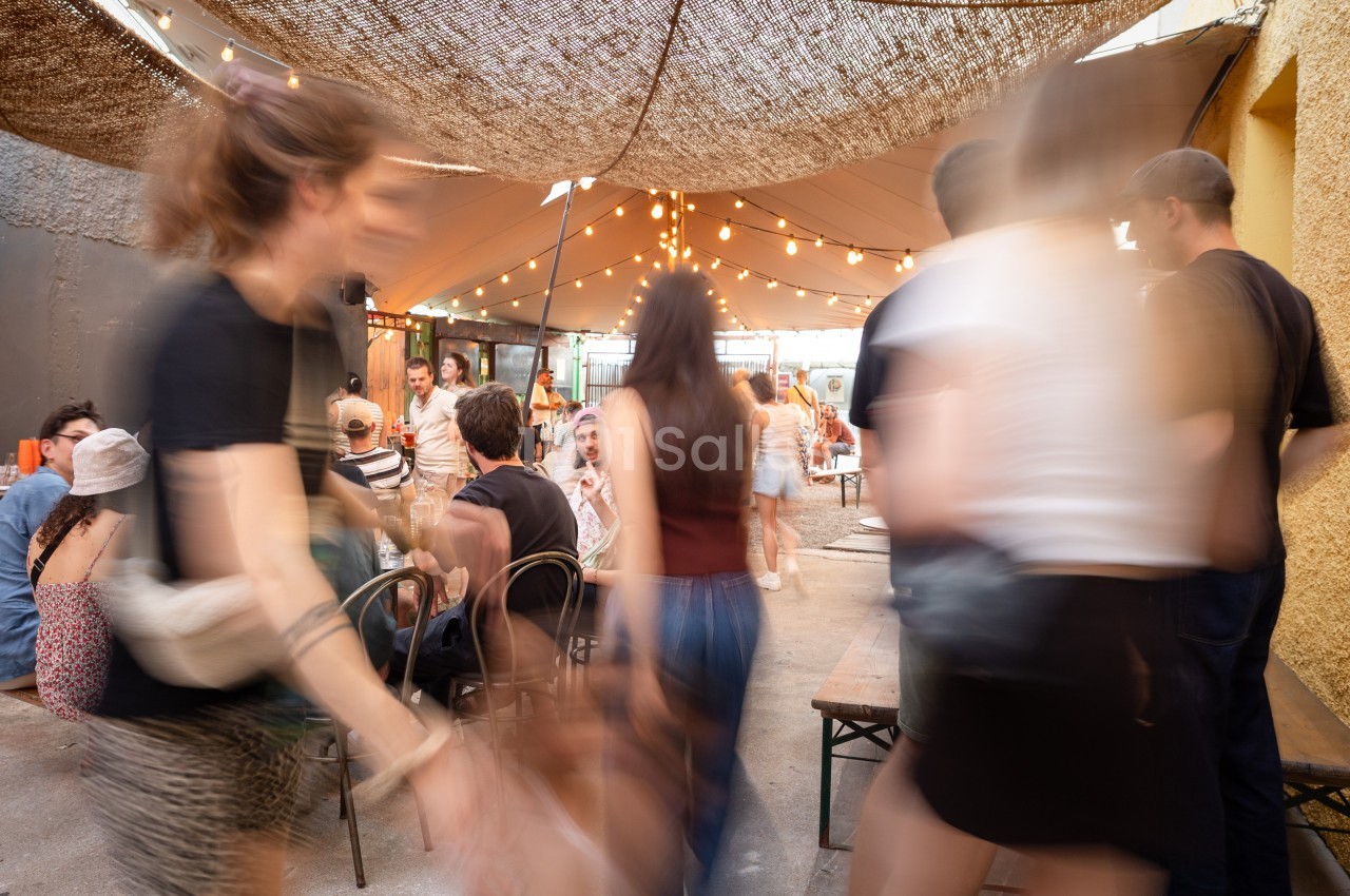 Personnes se déplaçant dans un espace extérieur animé, avec des tables, des chaises et des guirlandes lumineuses suspendues.
