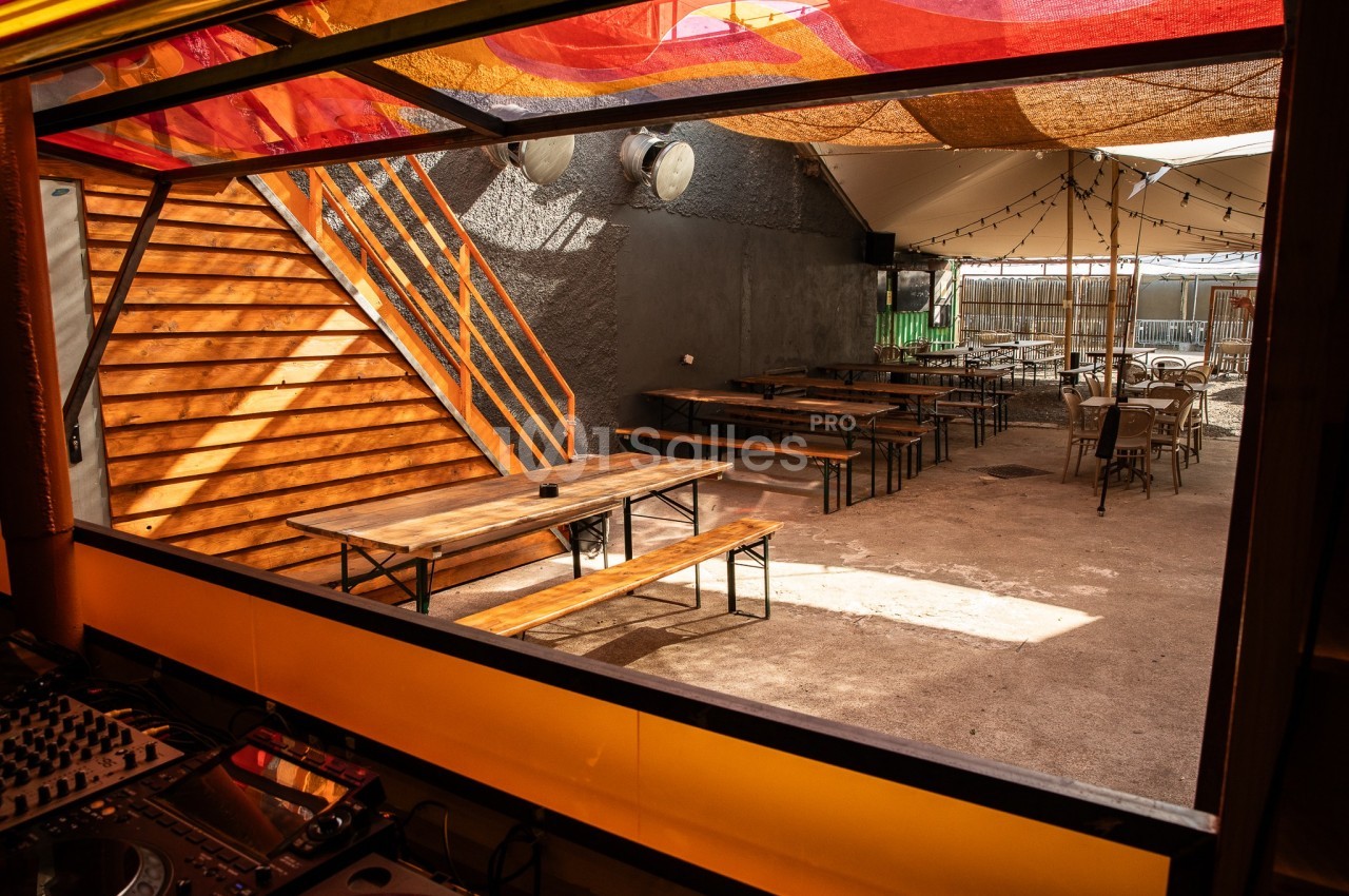 Terrasse extérieure avec tables en bois, bancs, escaliers en bois et toit partiellement couvert de toiles colorées.