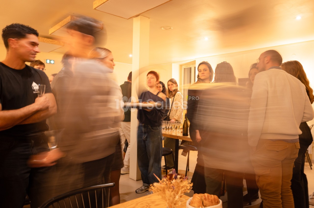 Groupe de personnes discutant dans une salle éclairée, ambiance conviviale avec des tables et des verres visibles.