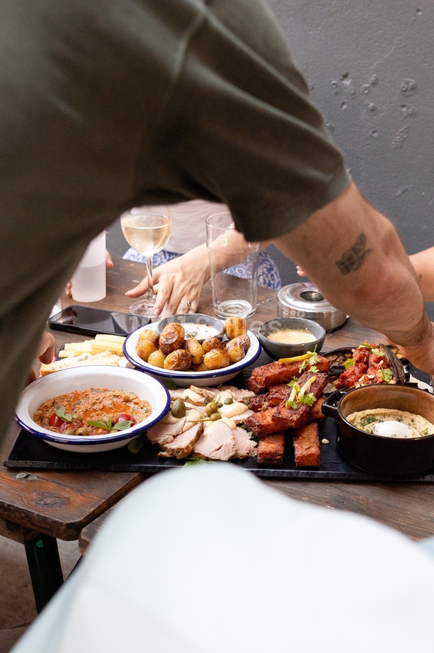 Un groupe partage un repas composé de viande, pommes de terre et accompagnements sur une table en extérieur.