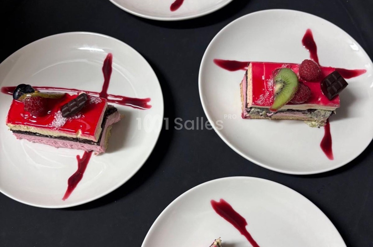 Assiettes contenant des parts de gâteau colorées, décorées de fruits, chocolat et coulis rouge sur fond noir.