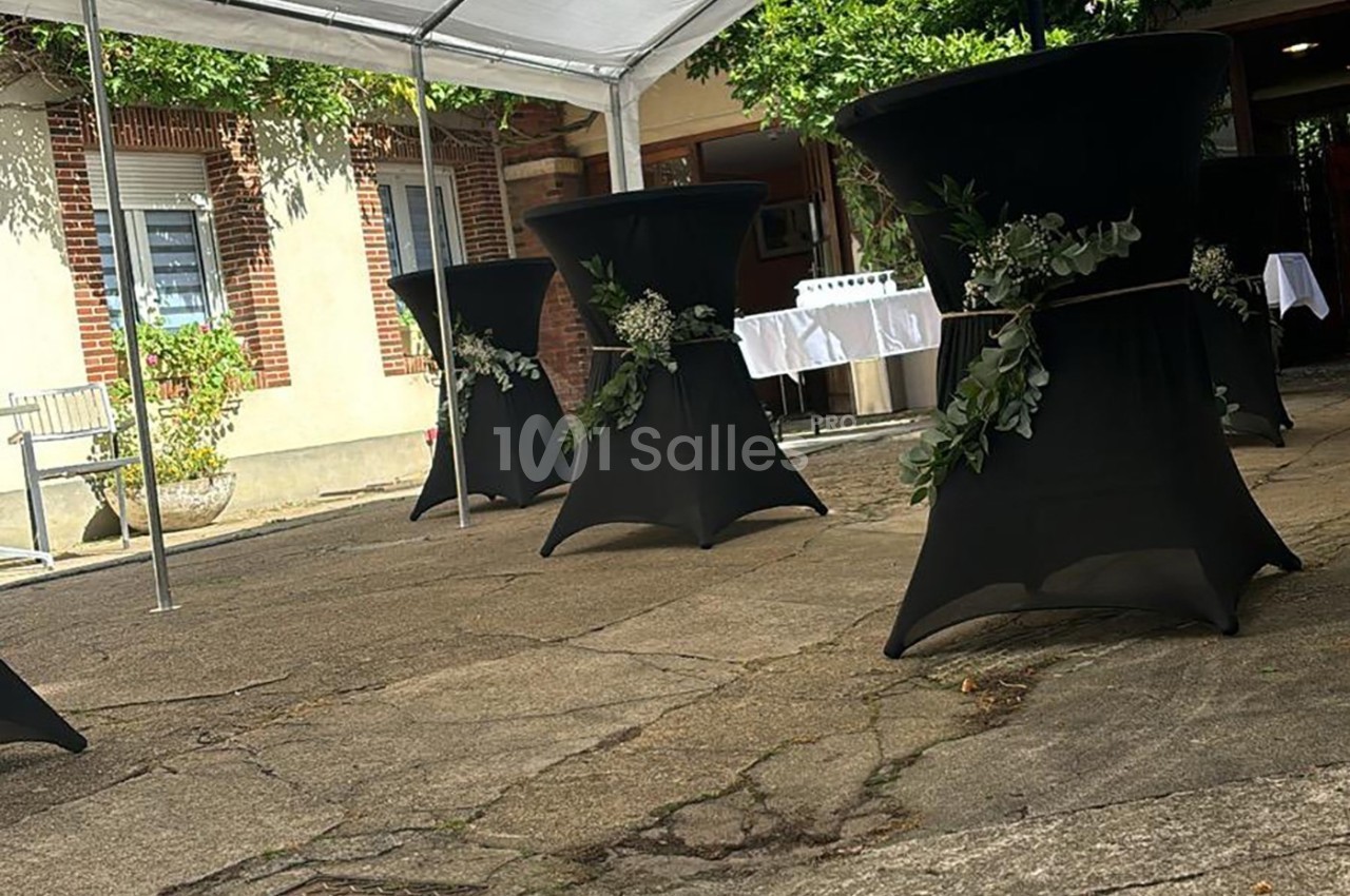 Tables hautes noires décorées de feuillages, disposées en extérieur sous une tente blanche, près d'un bâtiment en briques.
