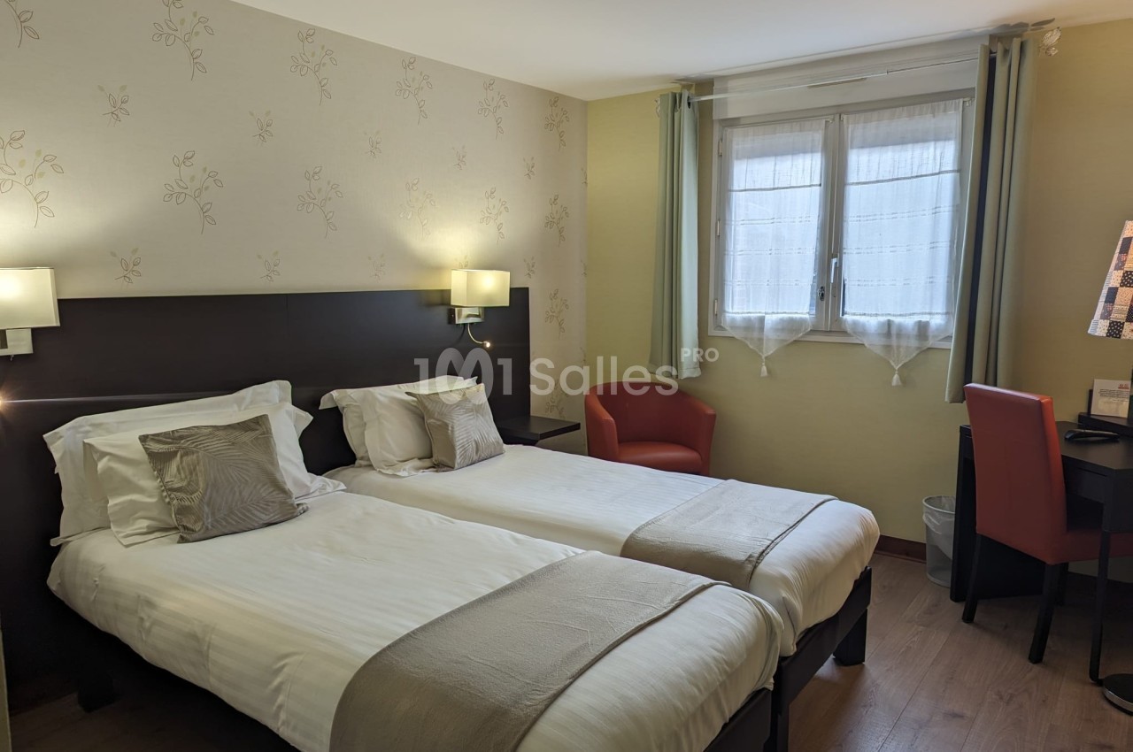 Chambre avec deux lits simples, lampes murales, bureau, chaise rouge et fenêtre avec rideaux translucides.