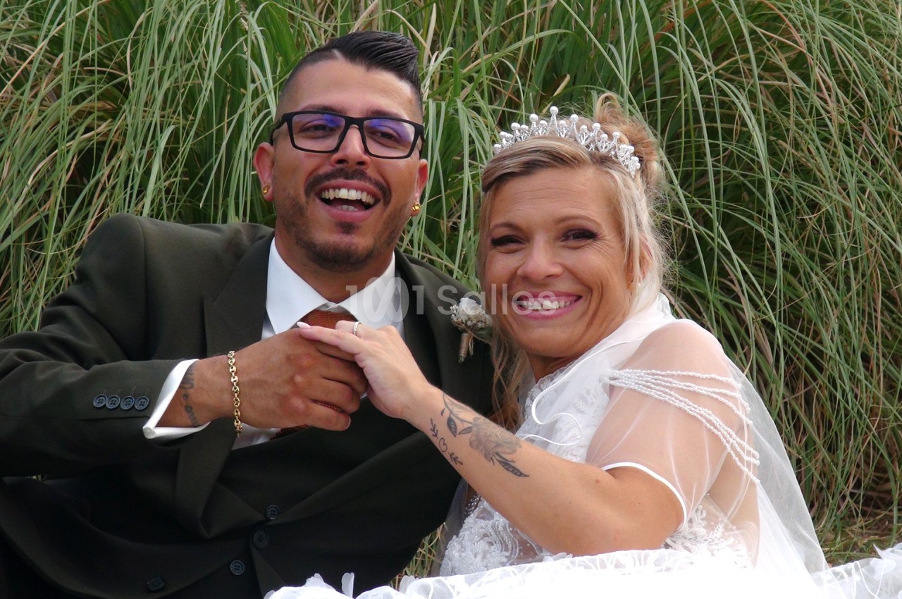 Un couple souriant en tenue de mariage pose devant des herbes hautes.