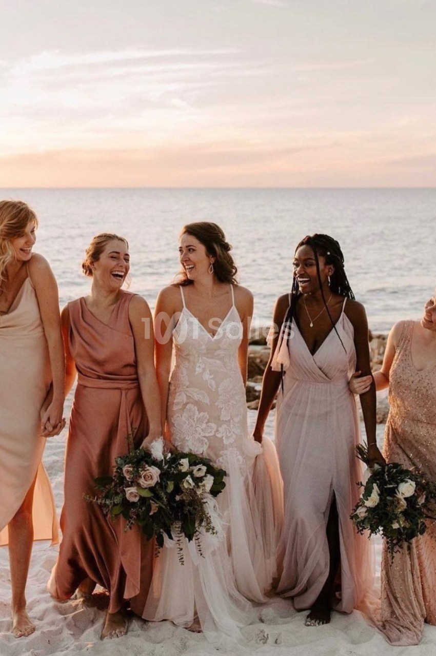 Cinq femmes en robes élégantes marchant sur une plage au coucher du soleil, tenant des bouquets de fleurs.
