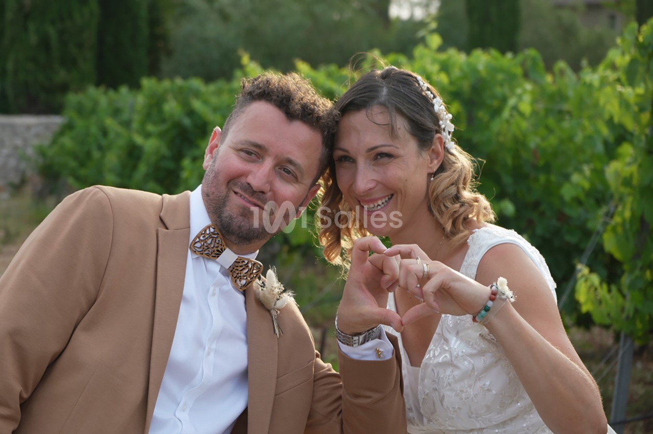 Un couple souriant en tenue de mariage formant un cœur avec leurs mains devant un décor naturel verdoyant.