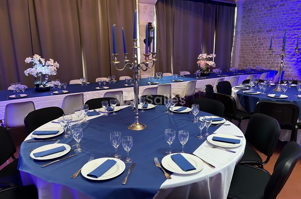 Salle décorée pour un événement avec tables rondes dressées, nappes bleues, chandeliers et éclairage tamisé.