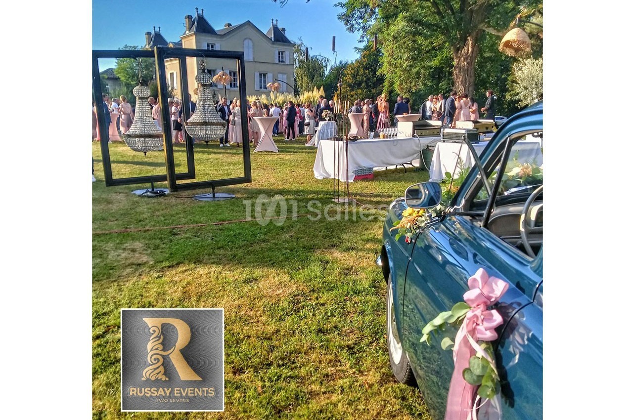 Vue d'un événement en plein air avec invités, décorations élégantes, voiture ancienne et un bâtiment en arrière-plan.