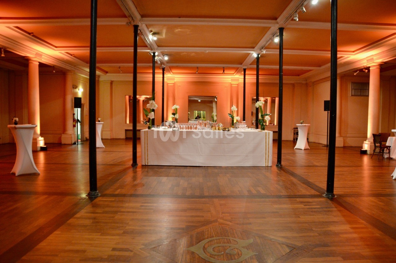 Salle de réception élégante avec parquet, colonnes blanches et table dressée pour un buffet au centre.
