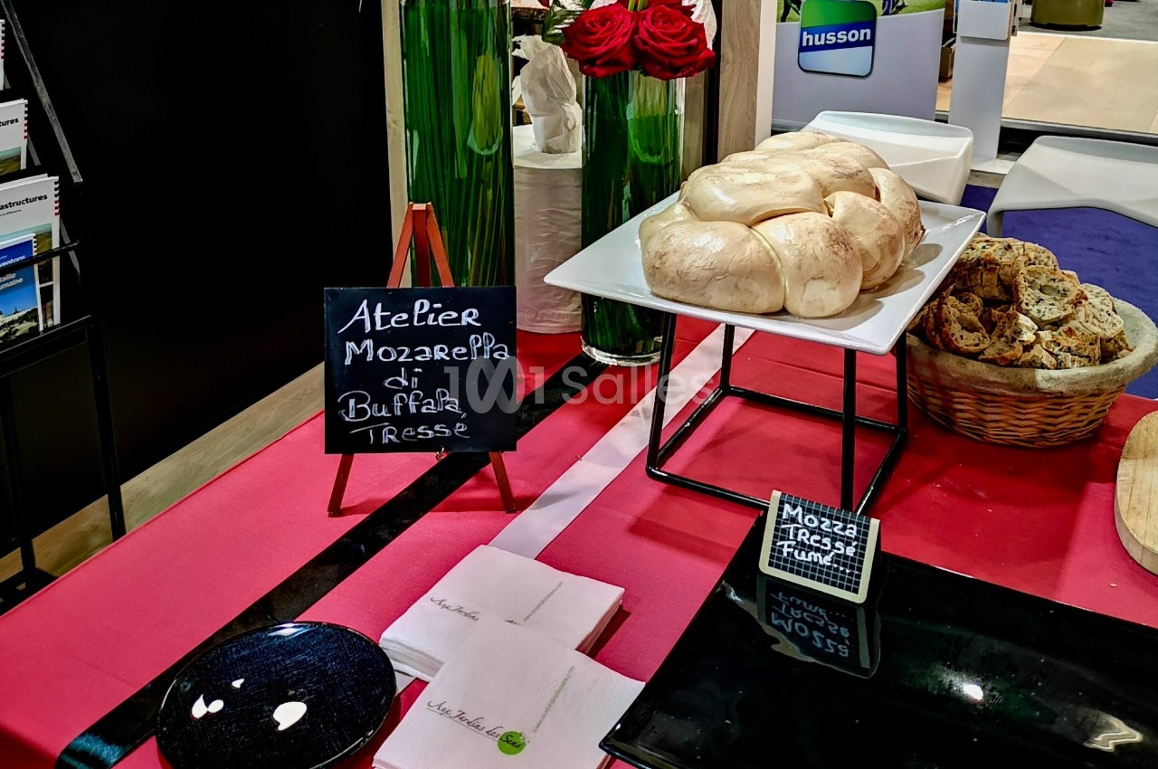 Plateau de mozzarella tressée sur une table rouge, accompagné de pain tranché et d'une ardoise indiquant ’Atelier Mozzarella…