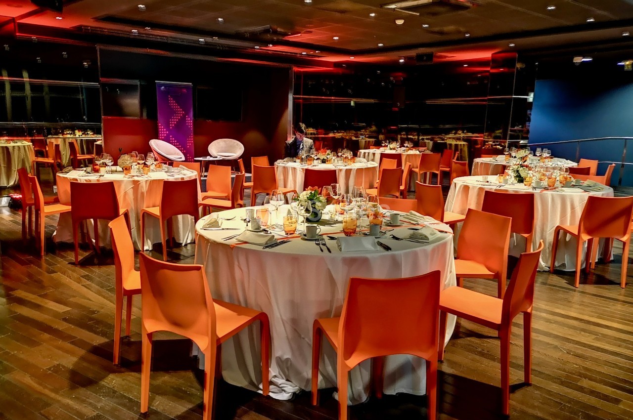 Salle de réception avec des tables rondes dressées, nappes blanches, chaises orange et éclairage tamisé.