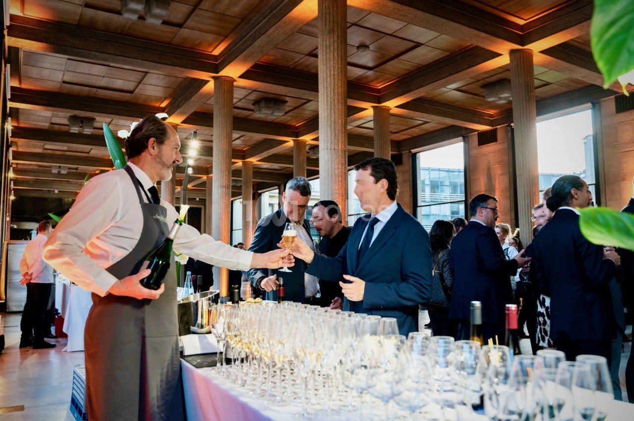 Un serveur sert du vin blanc à un homme lors d'un événement dans une salle élégante avec des invités en arrière-plan.