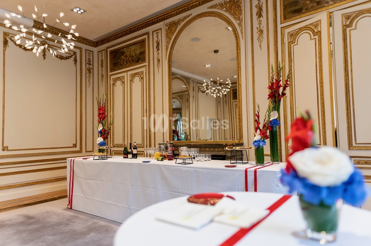 Salle élégante avec moulures dorées, table dressée avec boissons et décorations florales rouges, blanches et bleues.