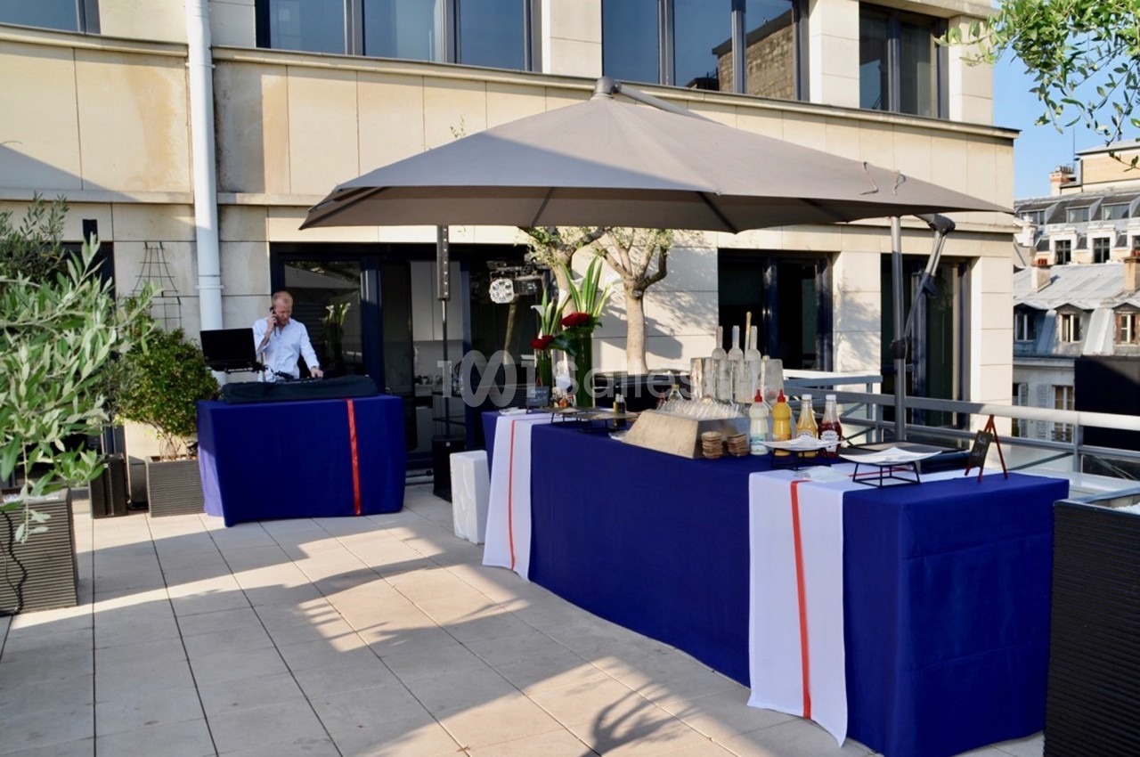 Buffet en extérieur sur une terrasse, avec nappes bleues et blanches, parasol et DJ en arrière-plan.