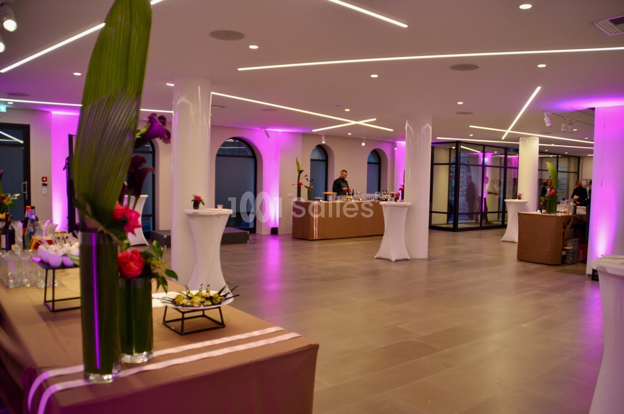 Salle de réception moderne avec éclairage rose, tables hautes et buffet décoré de fleurs.