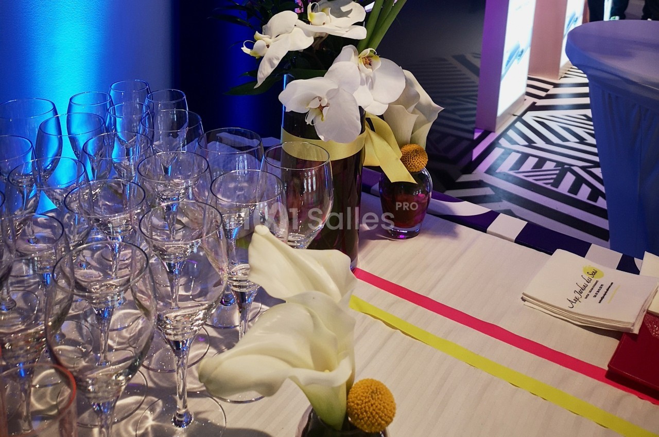 Verres vides alignés sur une table décorée de fleurs blanches et jaunes, éclairée par une lumière bleue.