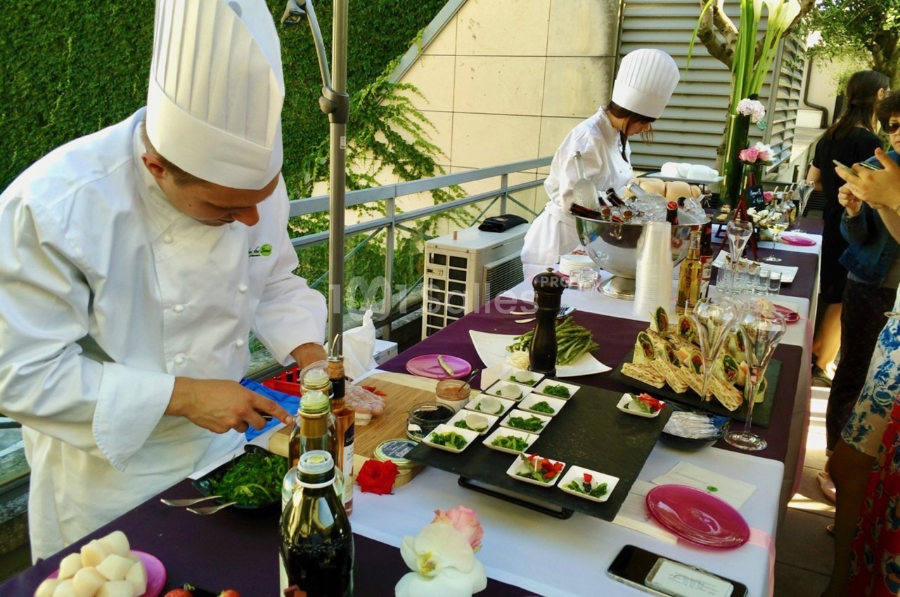 Un chef en tenue blanche prépare des plats sur une table garnie d'ingrédients et de décorations culinaires.