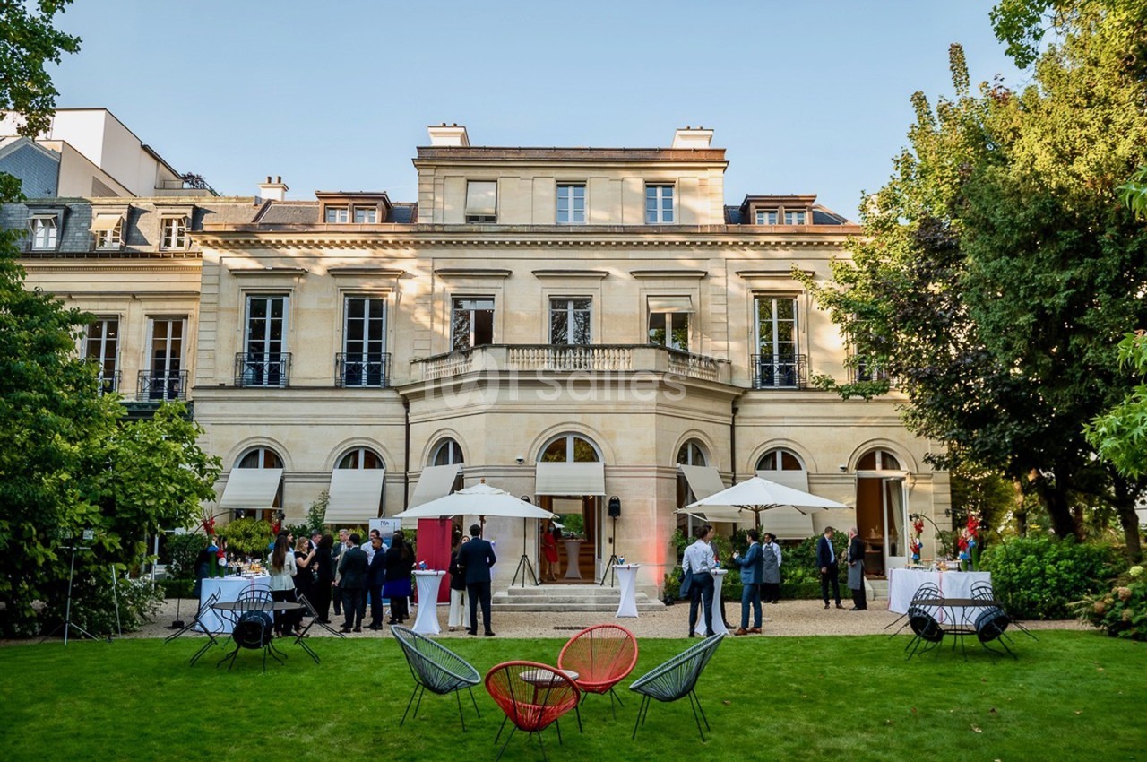 Façade d'un bâtiment élégant avec un jardin, des parasols et des personnes réunies lors d'un événement en plein air.