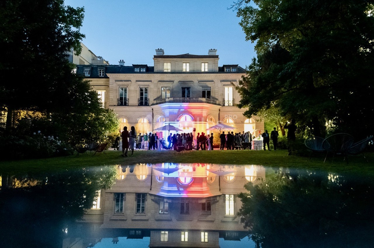 Façade illuminée d'un bâtiment élégant au crépuscule, avec un jardin et des invités rassemblés devant.