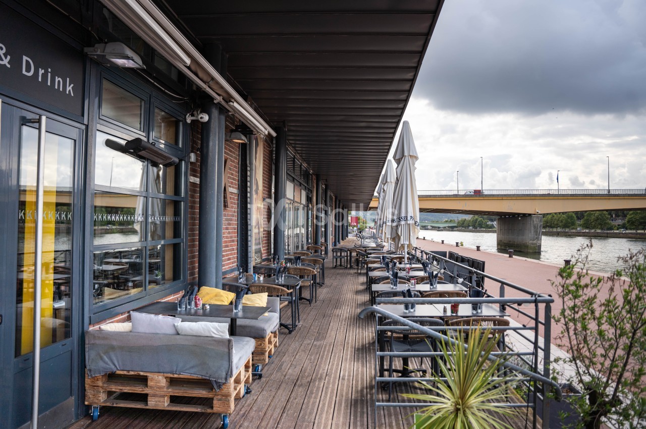 Terrasse en bois d'un restaurant au bord d'une rivière, avec tables, chaises et parasols sous un ciel nuageux.