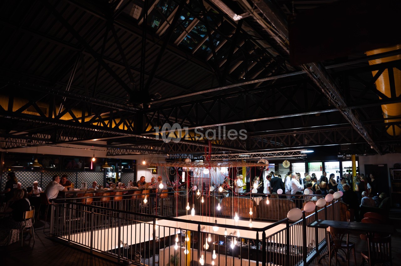 Intérieur d'un espace industriel aménagé en restaurant, avec des tables occupées et des lumières suspendues.