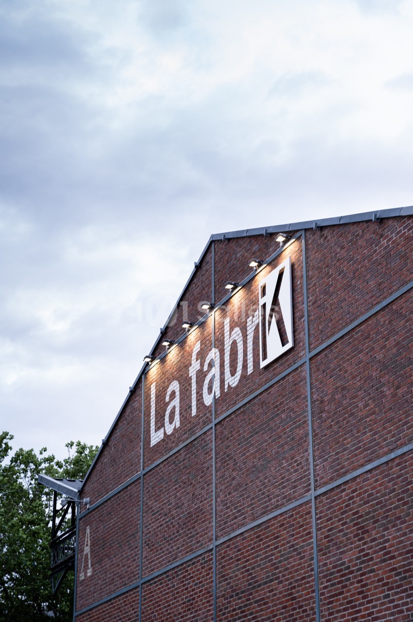 Façade en briques d'un bâtiment industriel avec l'inscription ’La Fabrik’ éclairée, sous un ciel nuageux.