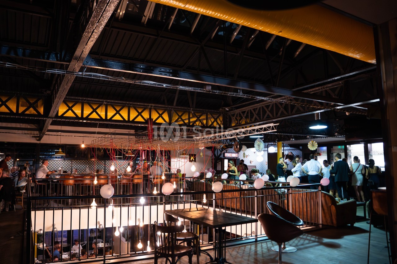 Espace intérieur industriel avec mezzanine, tables, chaises et éclairage tamisé, des personnes discutent en arrière-plan.