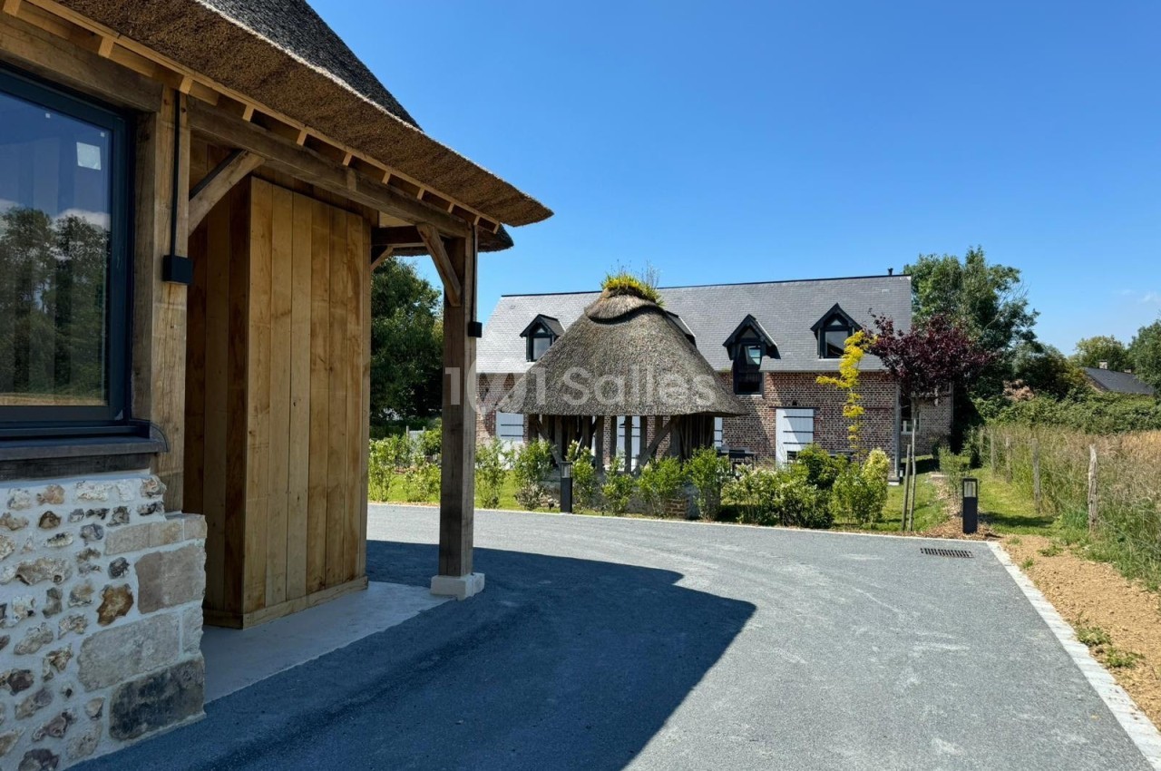 Maison en pierre et bois avec toit de chaume entourée de végétation, vue depuis une allée asphaltée.