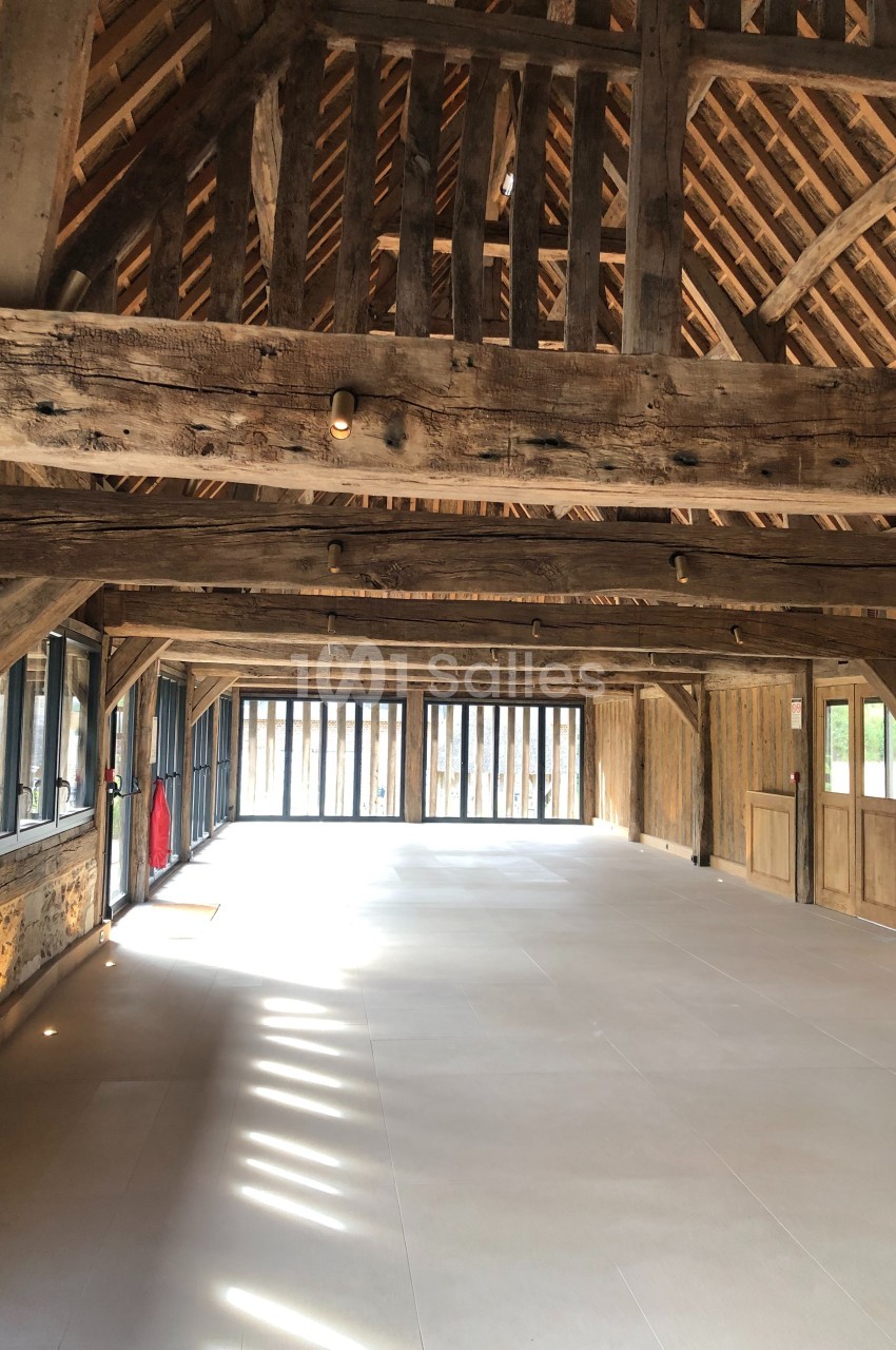 Intérieur d'une grande grange rénovée avec charpente en bois apparente et larges baies vitrées laissant entrer la lumière.