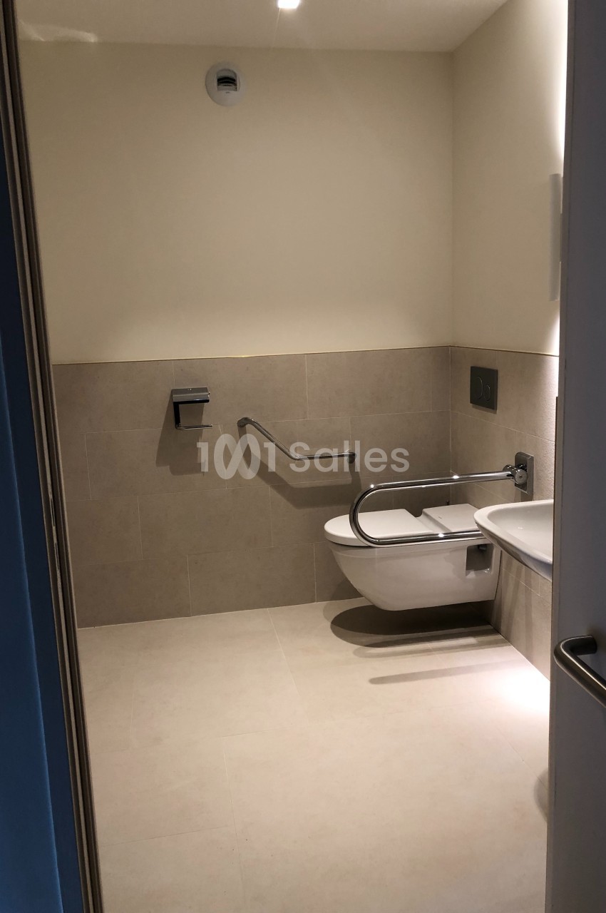 Toilettes accessibles avec barre d'appui, cuvette suspendue et distributeur de papier mural, éclairage sobre.