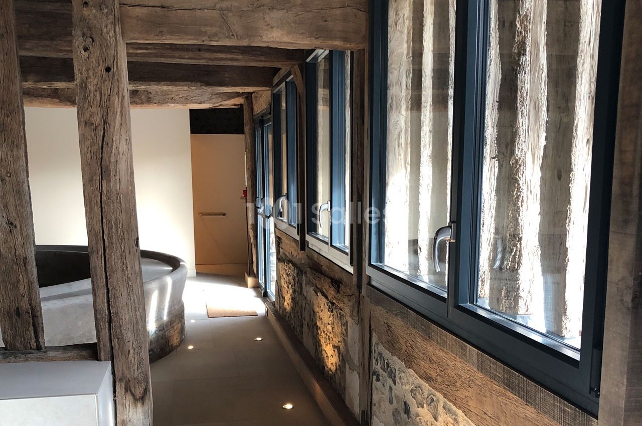 Vue intérieure d'un bâtiment avec des poutres en bois apparentes, des fenêtres modernes et un sol en pierre claire.