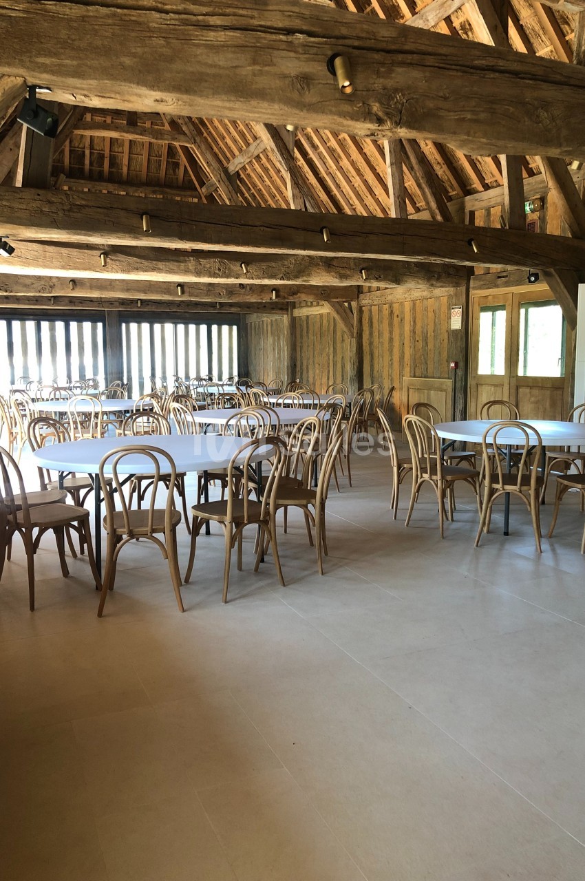 Salle avec charpente en bois apparente, tables et chaises en bois disposées dans un espace lumineux.