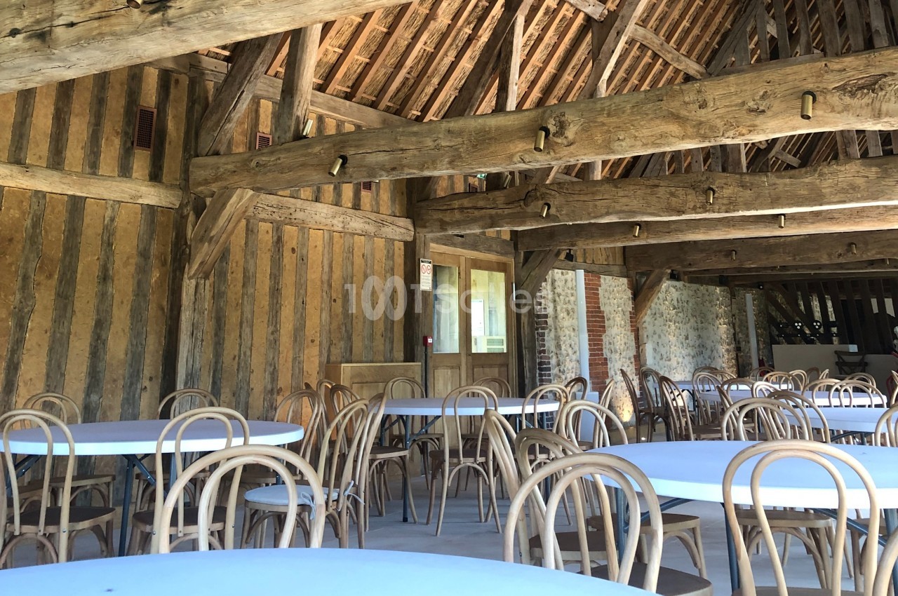 Salle avec charpente en bois apparente, tables rondes blanches et chaises en bois disposées pour un événement.