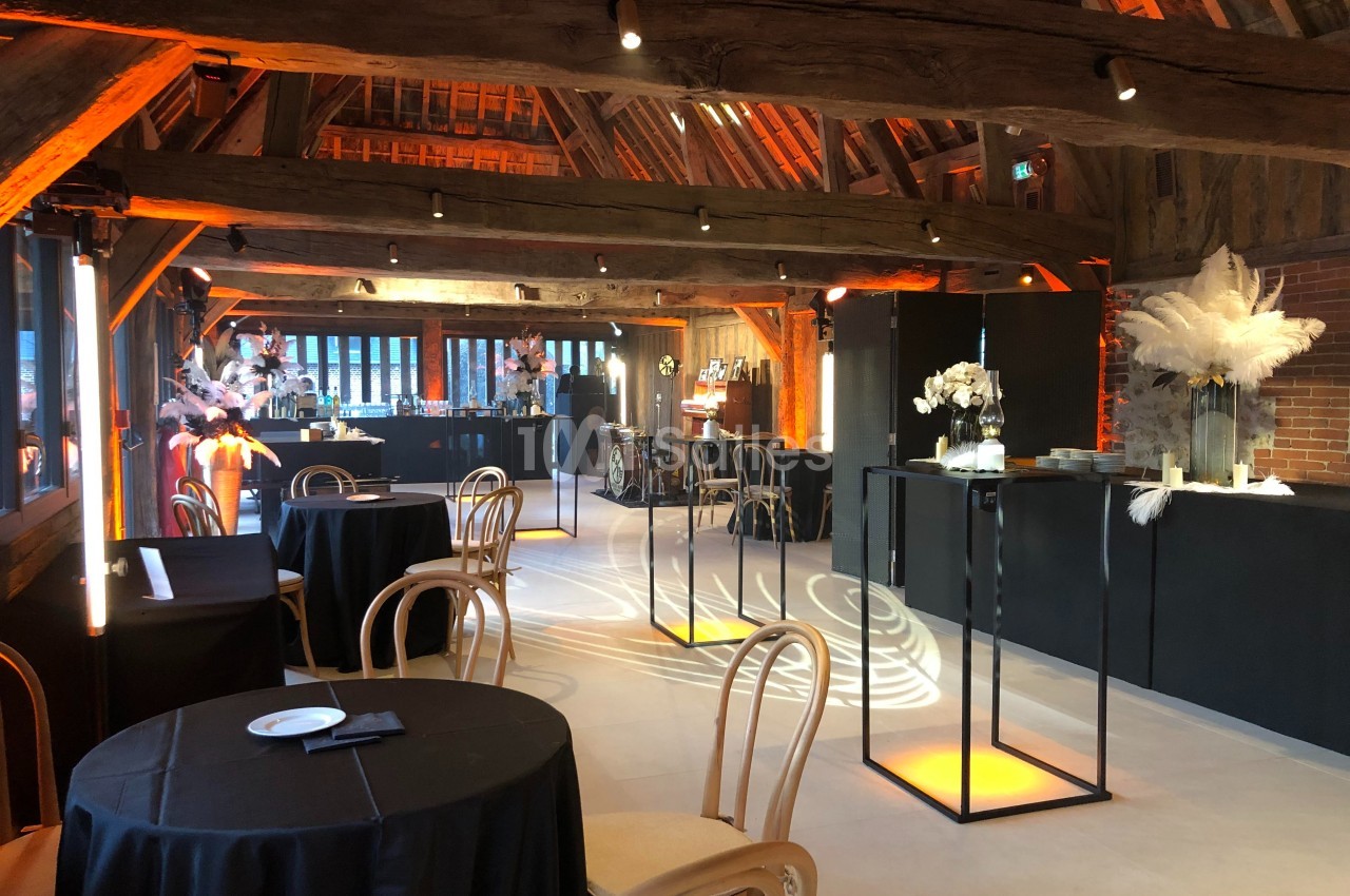 Salle de réception avec poutres apparentes, tables rondes, chaises en bois et décorations lumineuses élégantes.