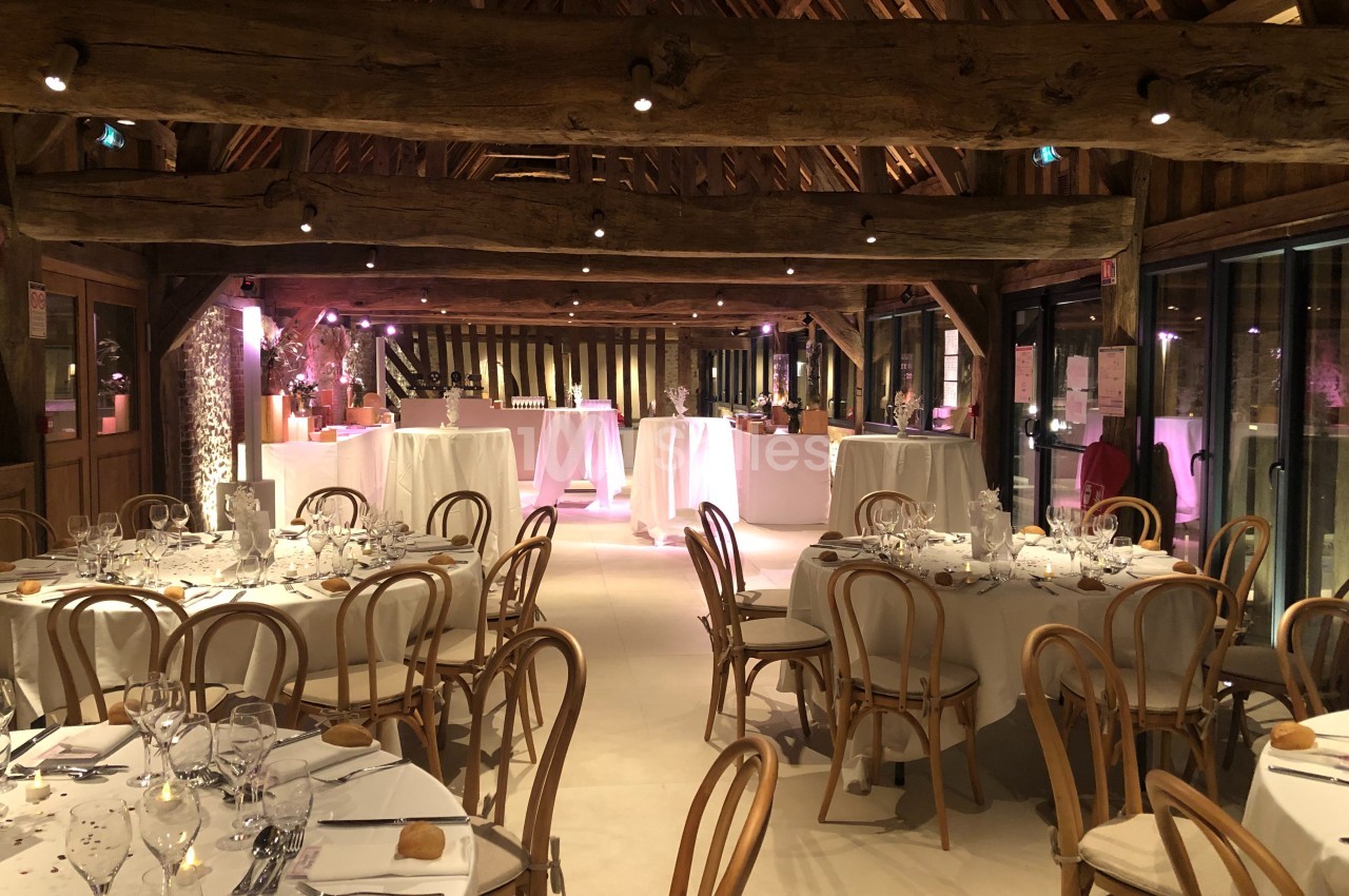Salle de réception avec tables rondes dressées, chaises en bois et éclairage chaleureux sous une charpente en bois apparente.