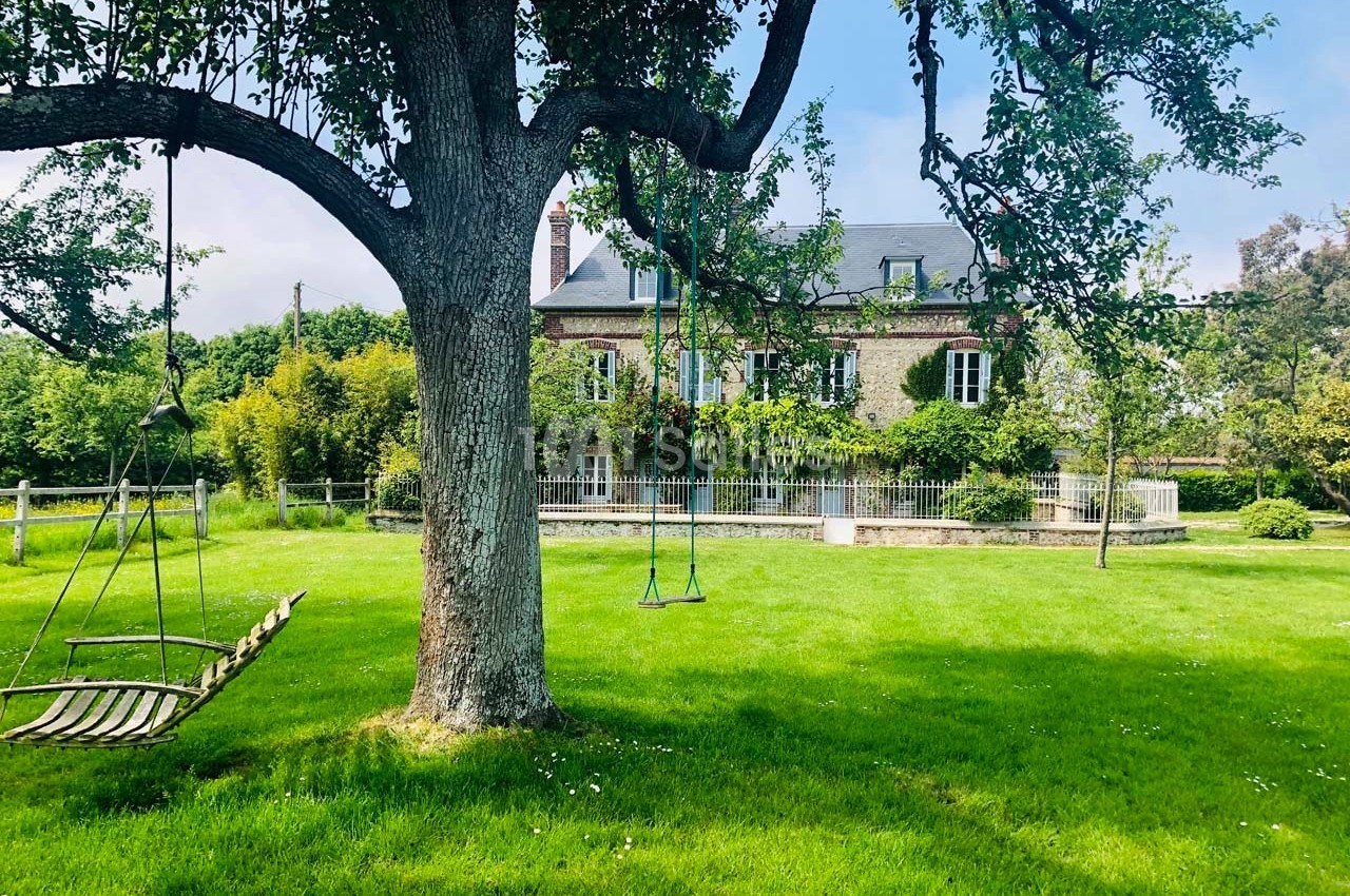 Balancelles suspendues à un arbre dans un jardin verdoyant, avec une maison en pierre en arrière-plan.