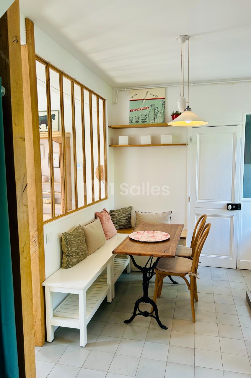 Coin repas lumineux avec table en bois, banc avec coussins, chaises, étagère murale et verrière intérieure.