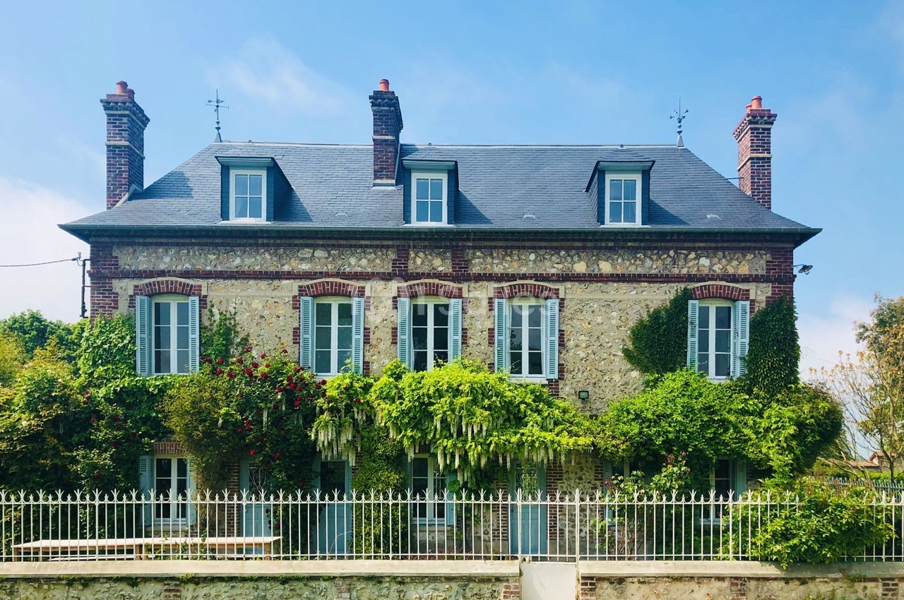 Façade d'une maison en pierre avec volets bleus, entourée de végétation et clôturée par une grille blanche.