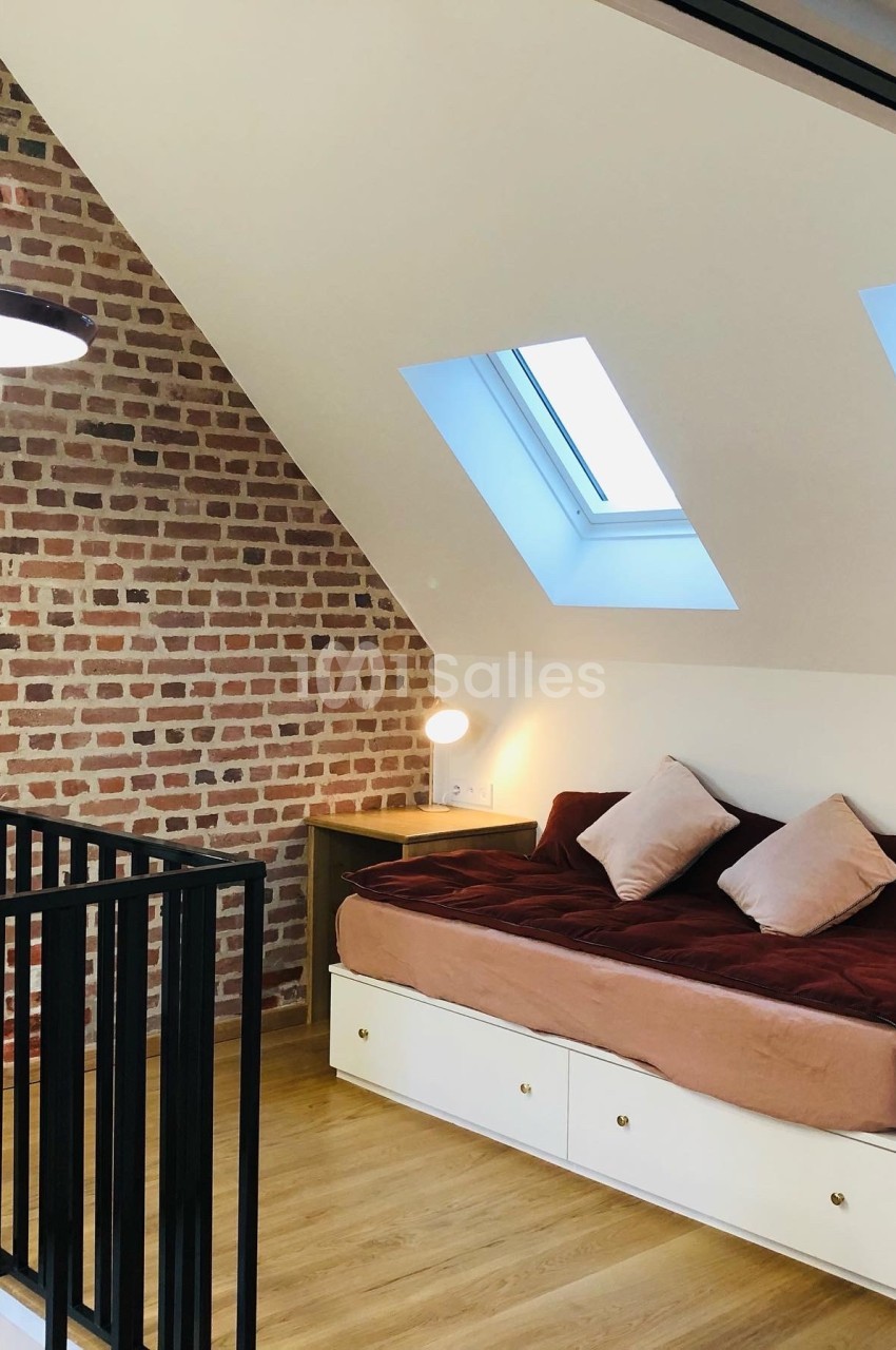 Chambre mansardée avec mur en briques, lit avec coussins, parquet clair et lumière naturelle provenant de deux fenêtres de…