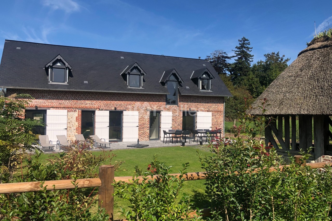 Maison en briques avec toit en ardoise, jardin aménagé et terrasse, entourée de verdure et d'une structure en chaume.