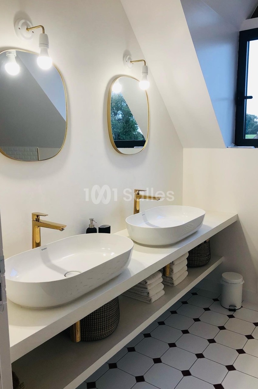 Salle de bain moderne avec deux vasques blanches, miroirs ovales dorés et éclairage mural sous un toit mansardé.