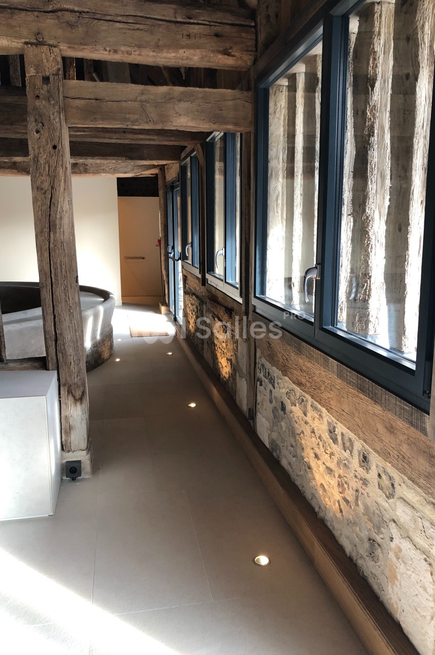 Couloir lumineux avec murs en pierre et poutres en bois apparentes, éclairé par des spots au sol et des fenêtres modernes.