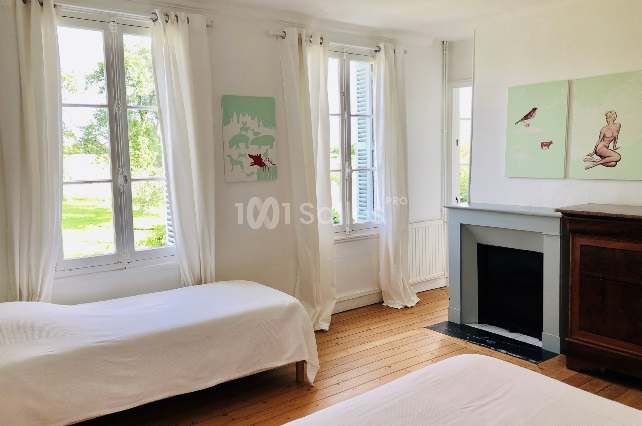 Chambre lumineuse avec deux lits simples, parquet en bois, cheminée décorative et grandes fenêtres avec rideaux blancs.