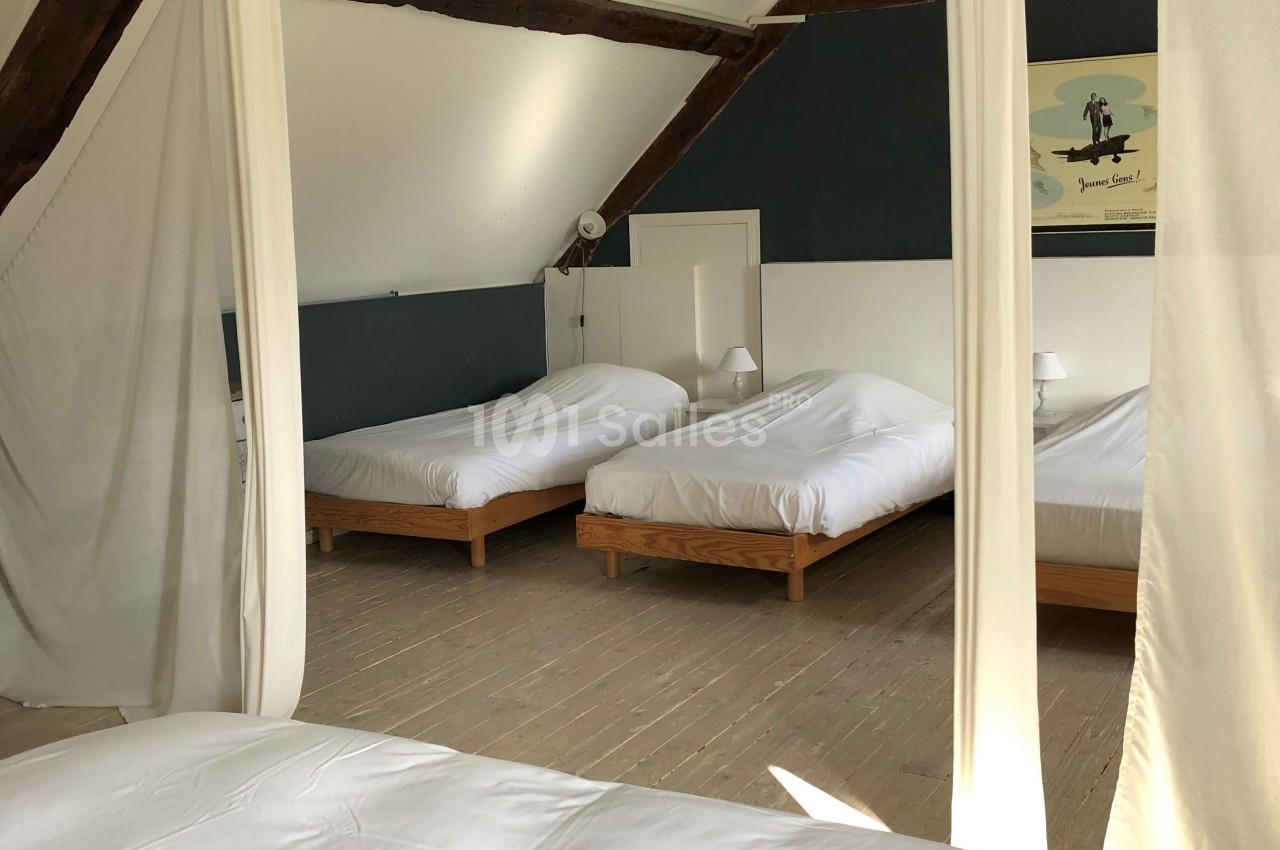 Trois lits simples avec draps blancs dans une chambre lumineuse aux poutres apparentes et parquet en bois clair.