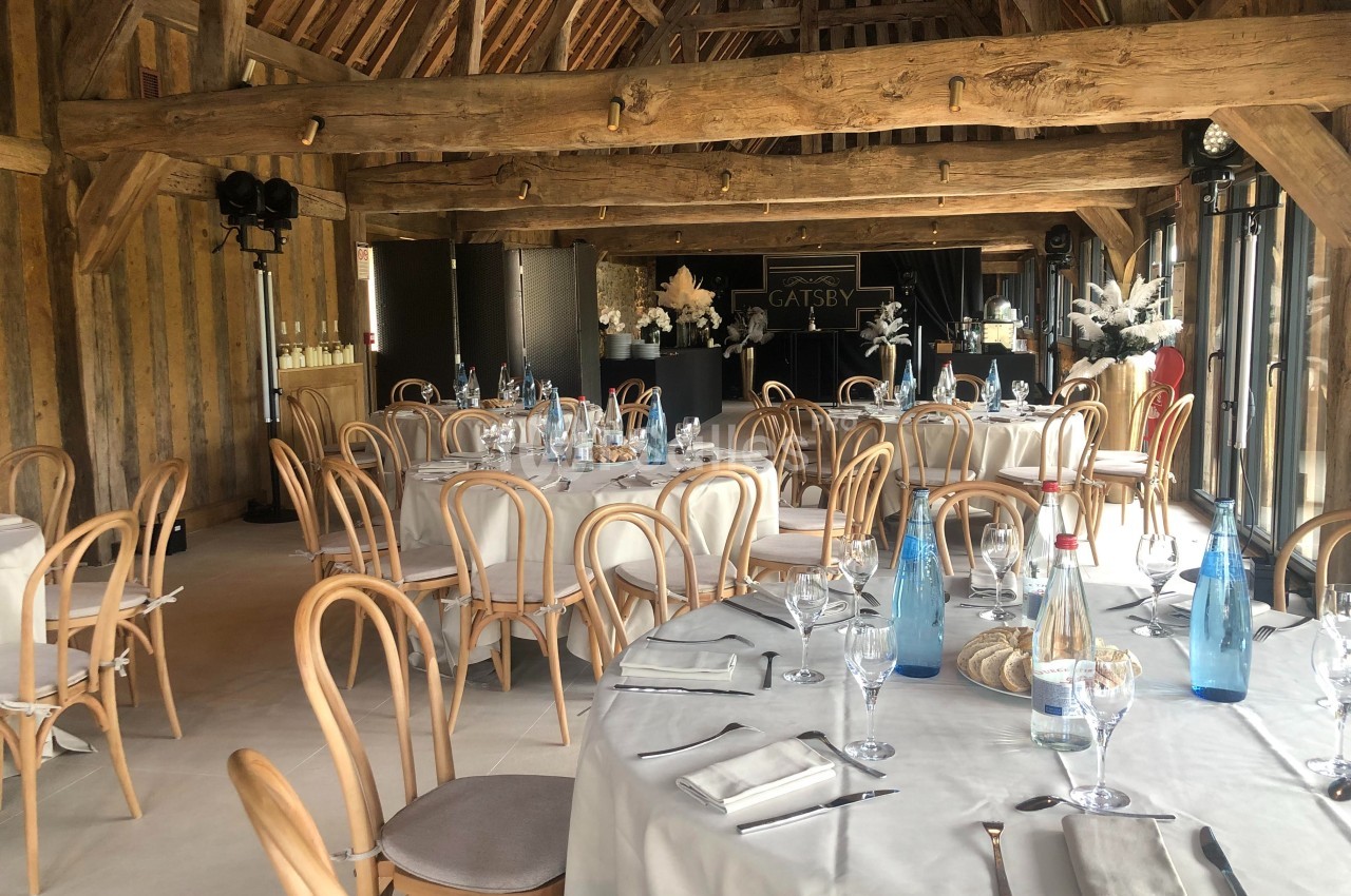 Salle de réception avec tables rondes dressées, chaises en bois clair et poutres apparentes sous un toit en bois.
